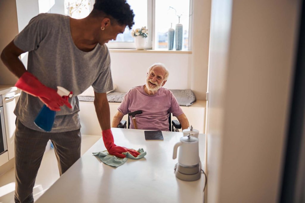 older person sitting in a wheelchair smiling at a care worker. Care worker is standing, cleaning a table.