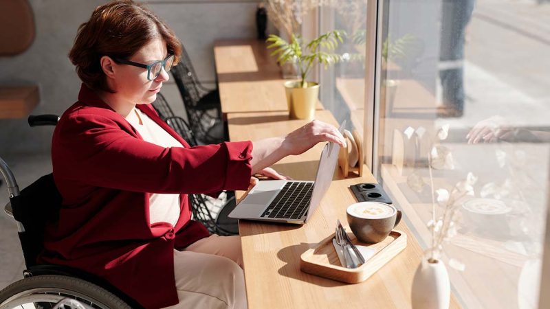 Image showing a woman who uses a wheelchair, sitting in a café, focusing on her laptop screen.