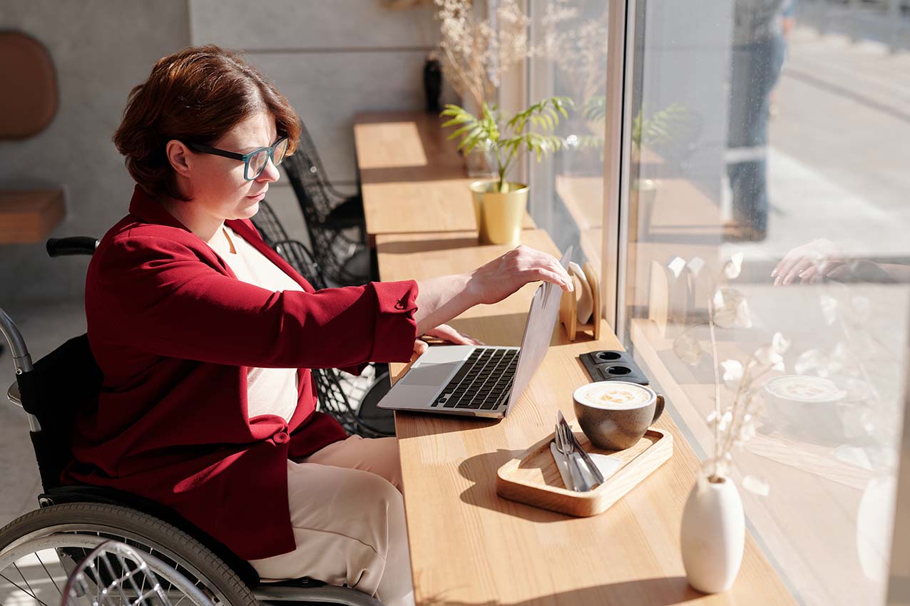 Image showing a woman who uses a wheelchair, sitting in a café, focusing on her laptop screen.